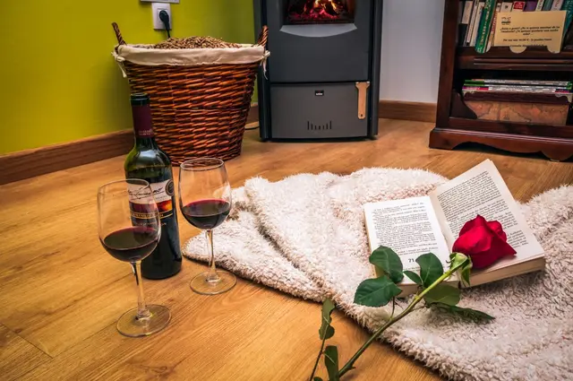 Suelo de madera con una manta blanca, dos copas de vino tinto, una botella de vino, un libro abierto con una rosa roja