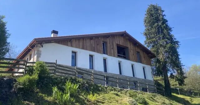 Vista exterior de una casa rural de dos plantas con fachada blanca y madera, tejado a dos aguas y chimenea, situada