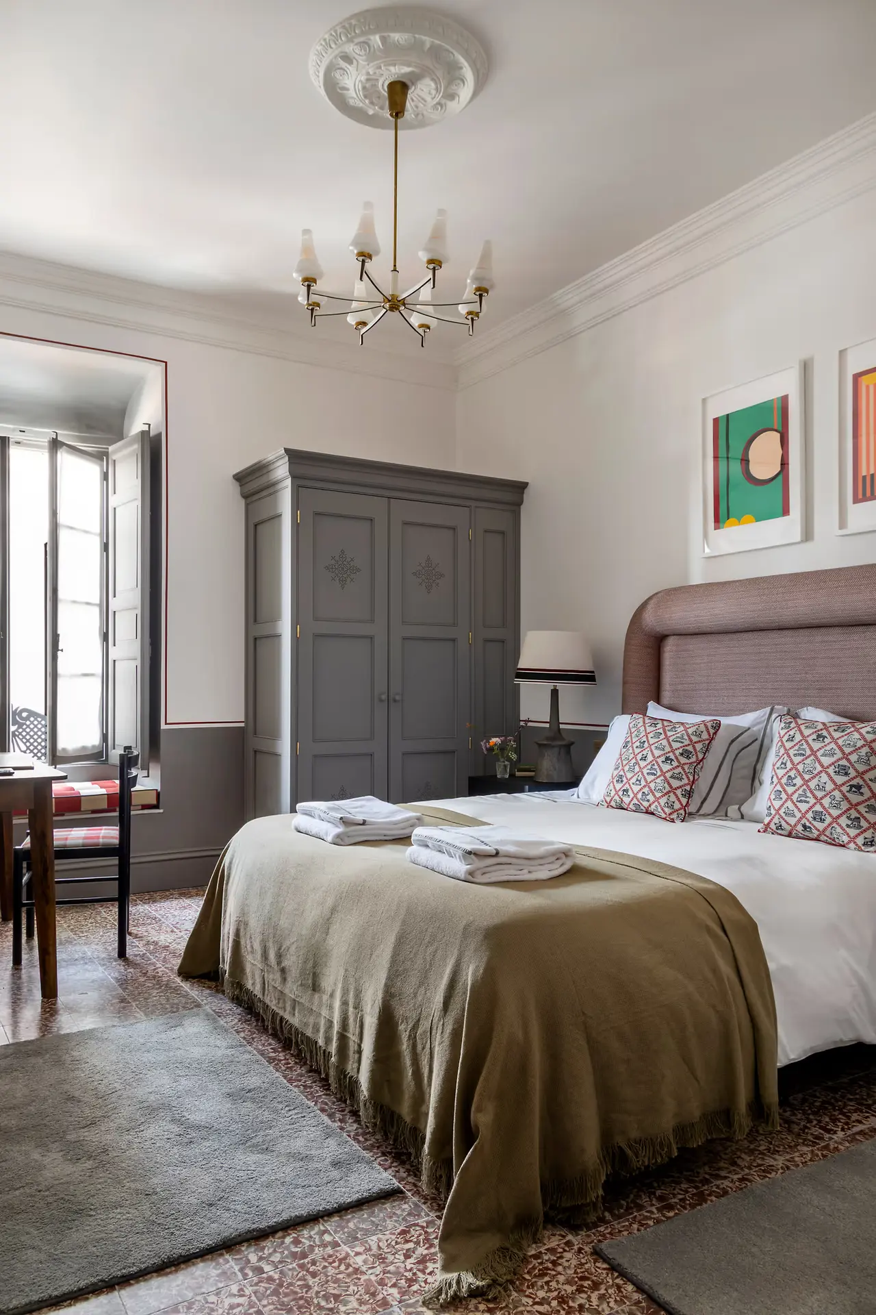 Habitación con cama grande vestida con colcha beige, armario gris, lámpara de araña en el techo, alfombras en el suelo