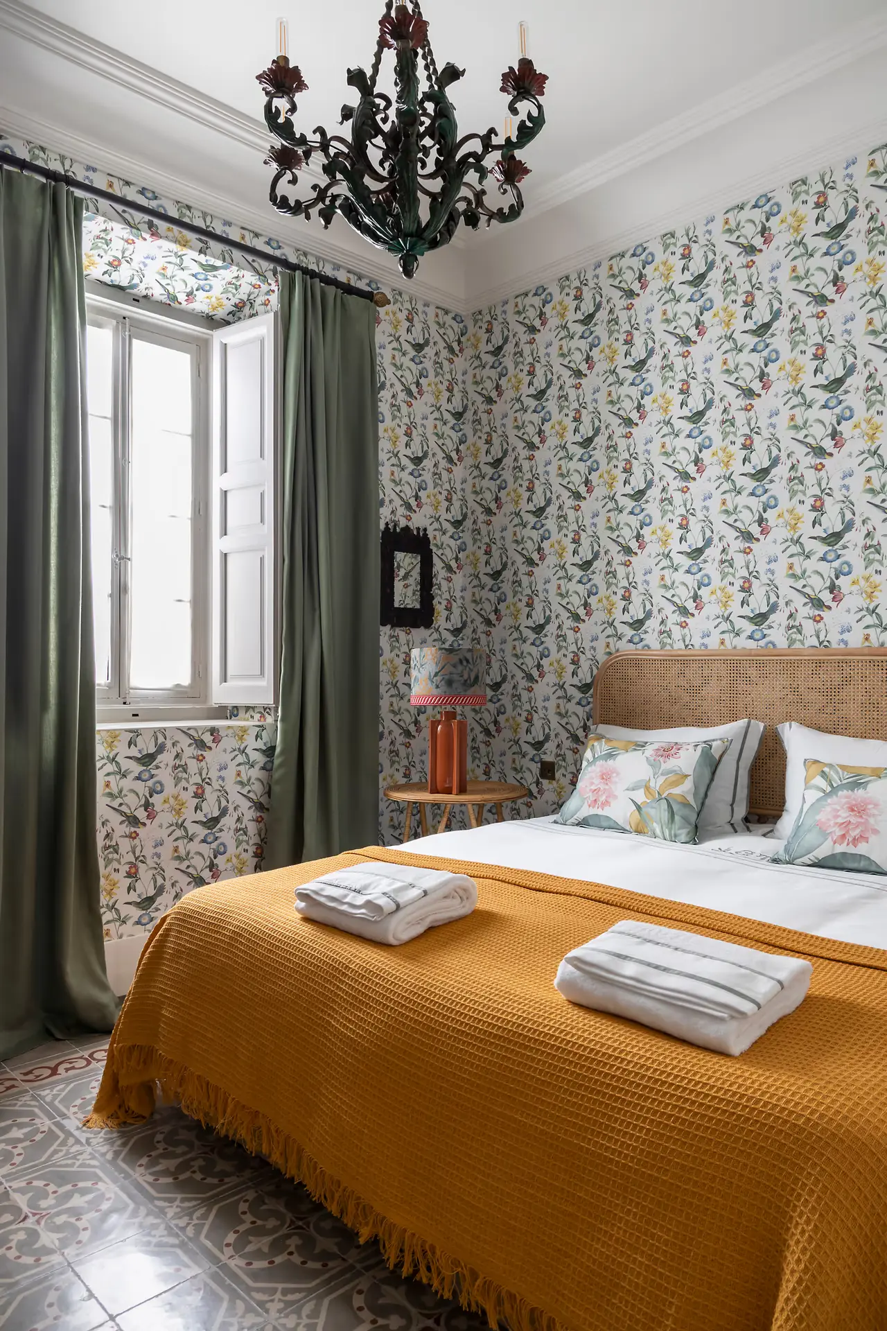 Habitación con cama grande cubierta por una colcha amarilla, papel pintado floral en las paredes, ventana con cortinas
