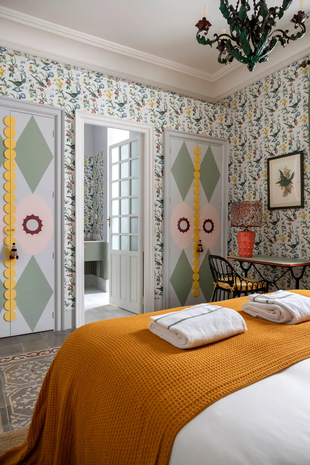 Habitación con cama vestida con colcha mostaza y toallas blancas dobladas, paredes con papel pintado floral, puertas
