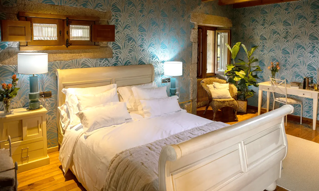 Habitación con cama grande blanca, cabecero iluminado, mesitas de noche con lámparas, papel pintado azul con motivos