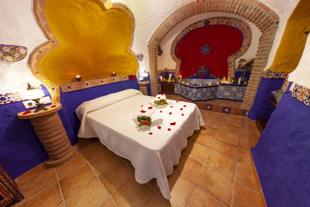 Habitación de cueva con cama doble vestida de blanco y pétalos de rosa, cabecero azul y amarillo, y al fondo