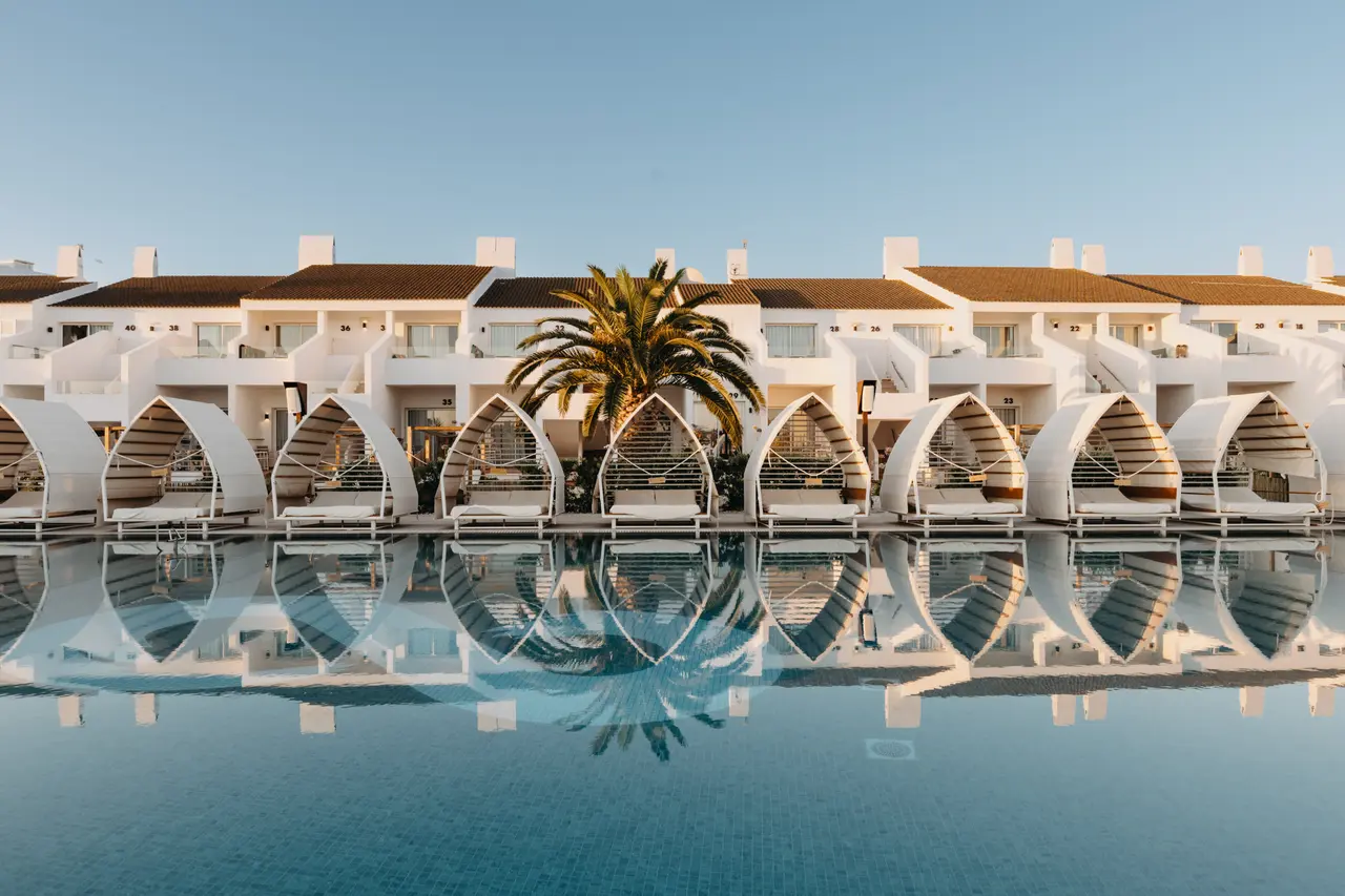 Vista de una piscina reflectante con palmera y edificios blancos de alojamiento al fondo bajo un cielo despejado.
