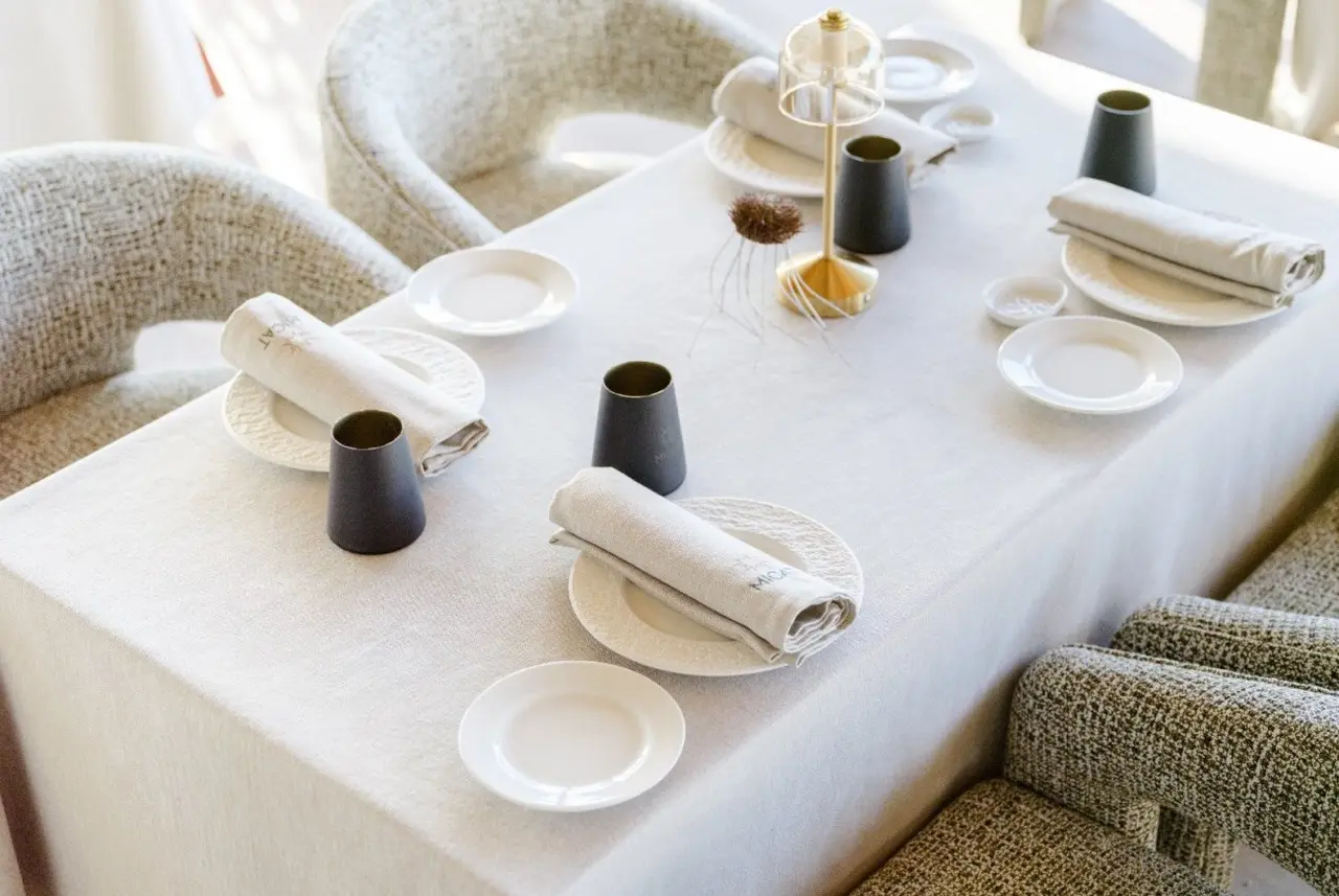 Mesa rectangular para cuatro comensales con mantel blanco, vajilla, servilletas enrolladas y copas negras, rodeada