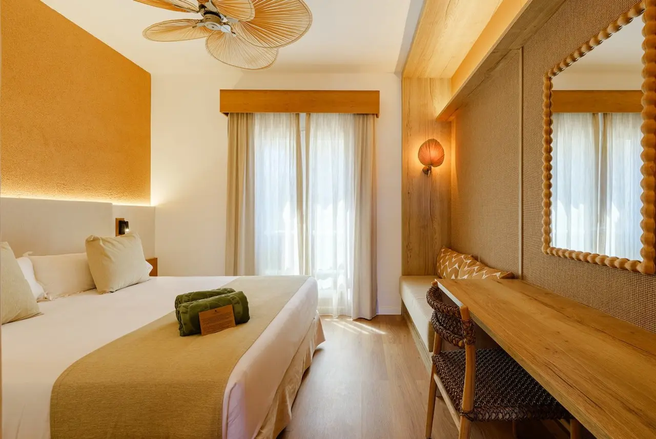 Habitación de hotel con cama grande, escritorio de madera, silla, ventana con cortinas y ventilador de techo.