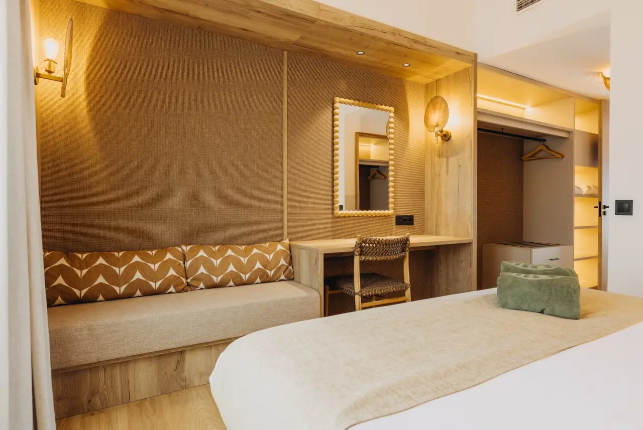 Habitación con cama, sofá, escritorio, espejo y armario empotrado en madera clara y tonos cálidos.