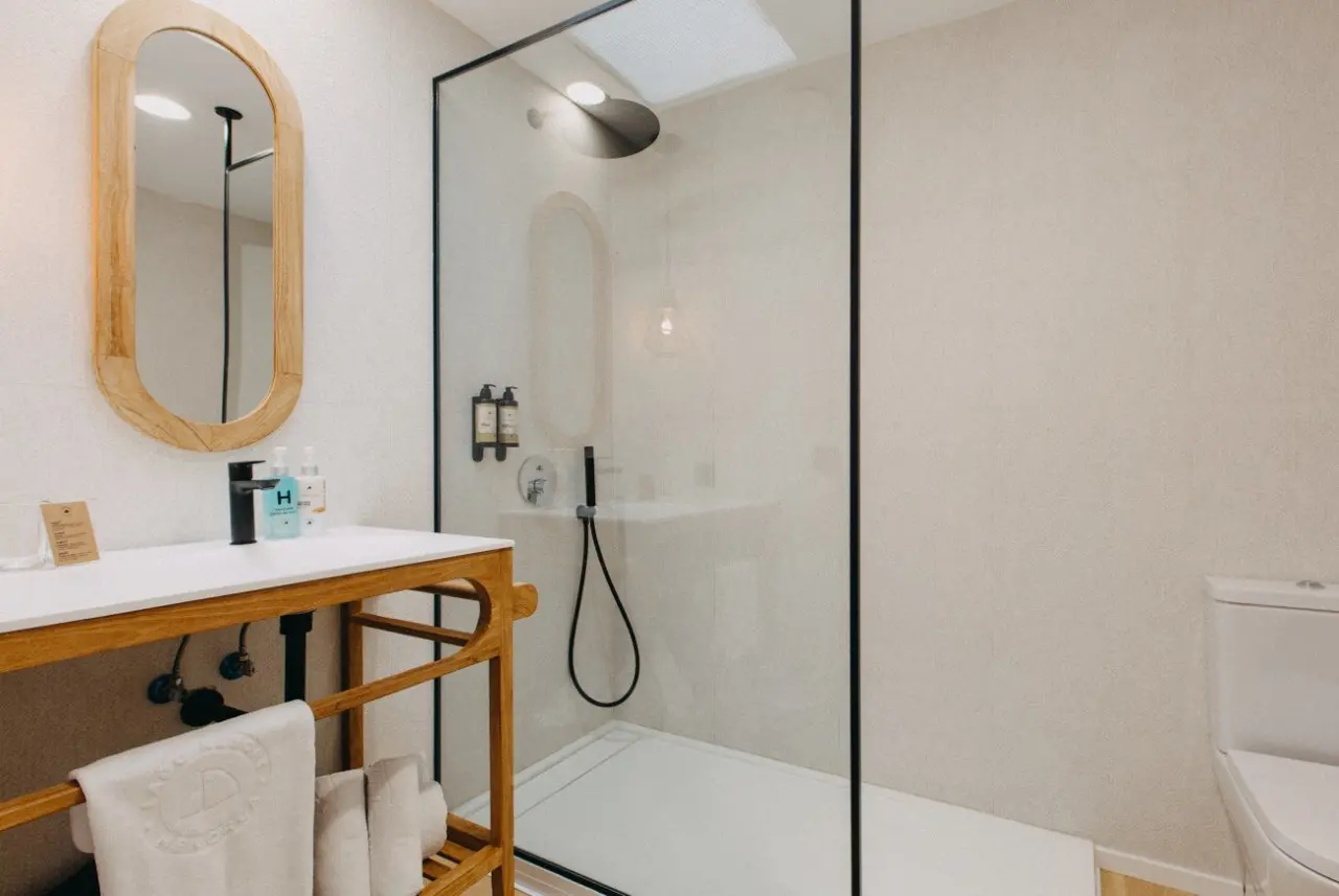 Baño con ducha de vidrio, lavabo de madera con espejo ovalado y toallas blancas.