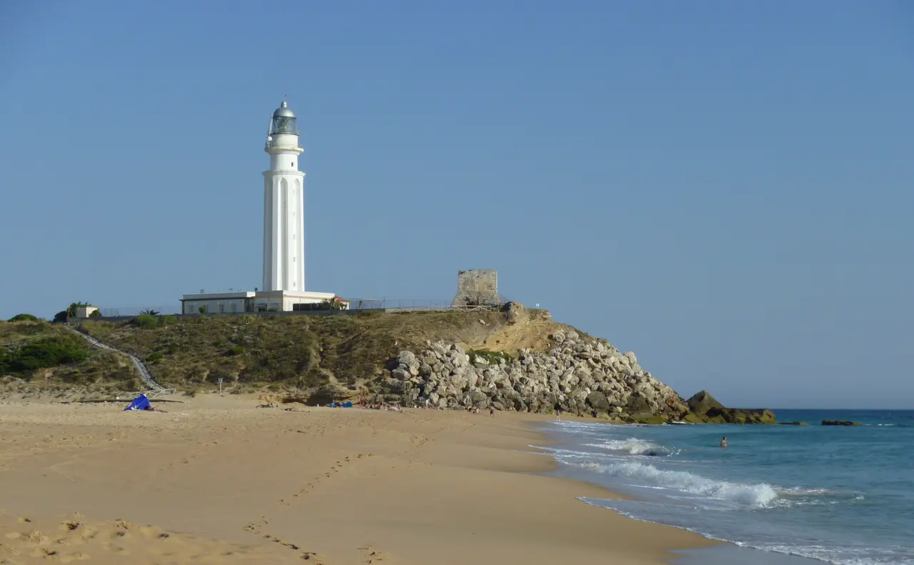 Vista de una playa de arena dorada con olas suaves, un faro blanco alto sobre un acantilado rocoso y un cielo azul