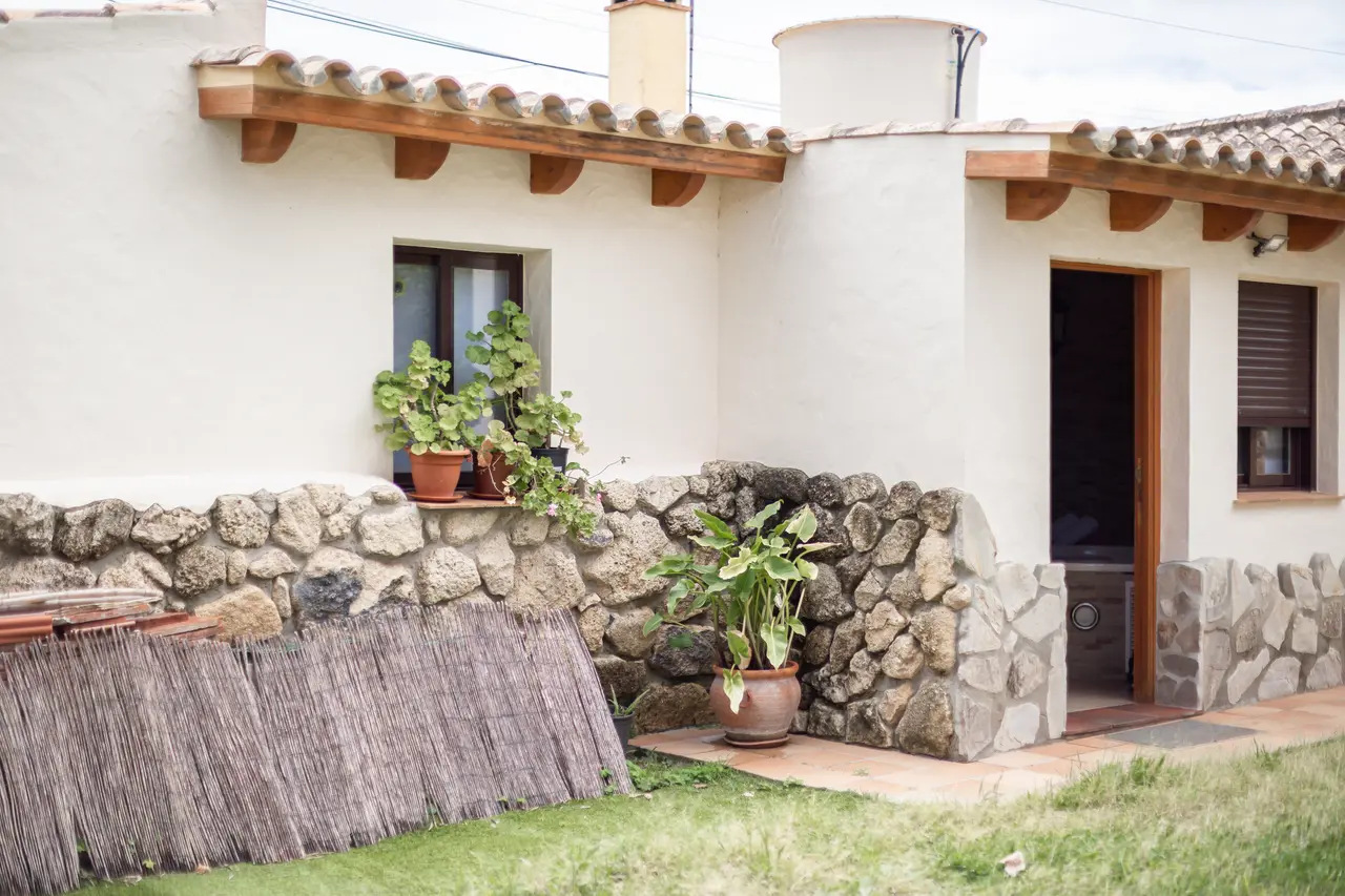 Fachada exterior de una casa blanca con techo de tejas, alféizar de piedra, macetas con plantas, puerta de entrada