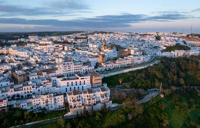 Vista panorámica de una ciudad blanca con edificios densos, torres y murallas, rodeada de vegetación y colinas