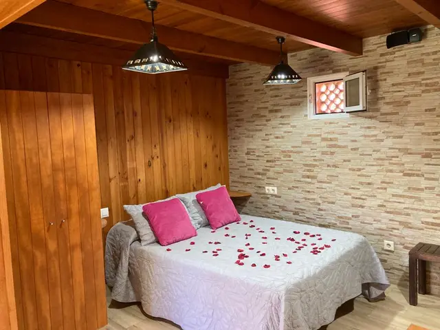 Habitación interior con paredes de madera y piedra, techo con vigas de madera, cama doble con colcha blanca y pétalos