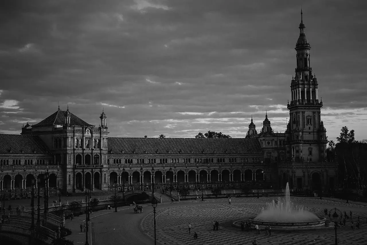 Vista en blanco y negro de una gran plaza con fuente central, edificios históricos con arcos y una torre alta al fondo