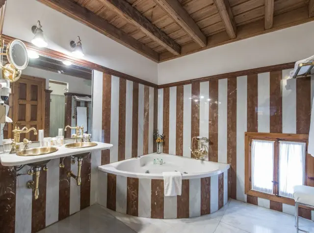 Jacuzzi triangular blanco empotrado en una esquina de un baño con paredes de mármol a rayas verticales marrones