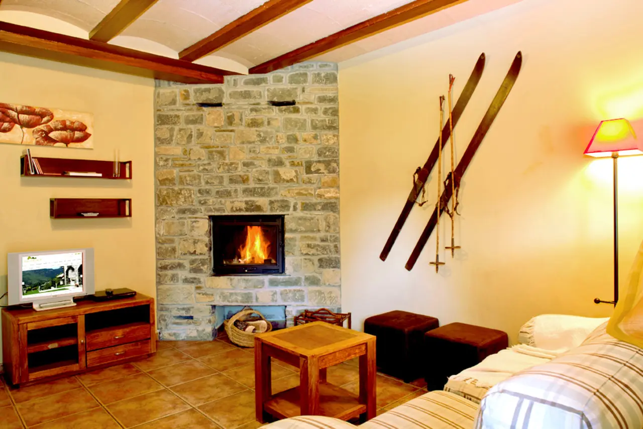 Interior de una sala con chimenea de piedra encendida, vigas de madera en el techo, televisión sobre un mueble
