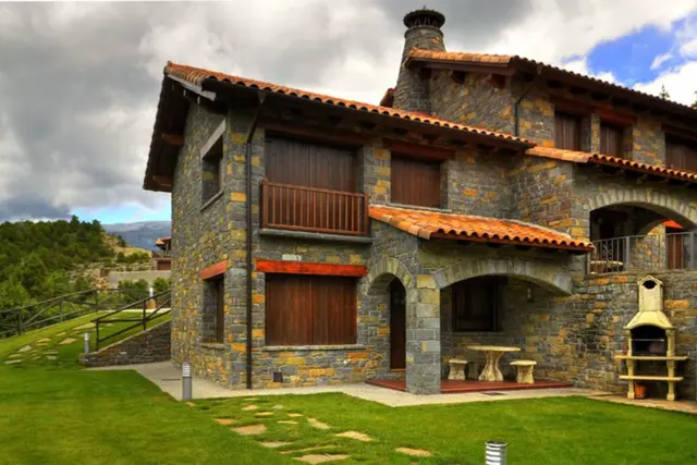 Vista exterior de una casa de piedra de dos plantas con tejado de teja, chimenea, balcones de madera y un patio