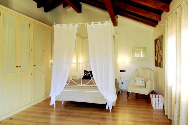Habitación interior con cama de dosel blanca, armario empotrado de color crema, butaca junto a la ventana y vigas