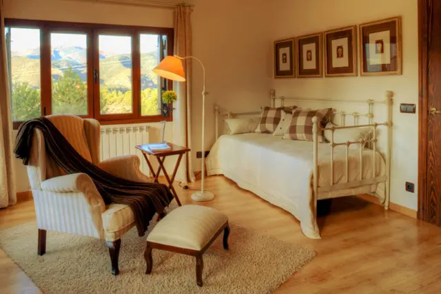 Habitación interior con cama de matrimonio, sillón orejero con reposapiés, ventana grande con vistas a montañas