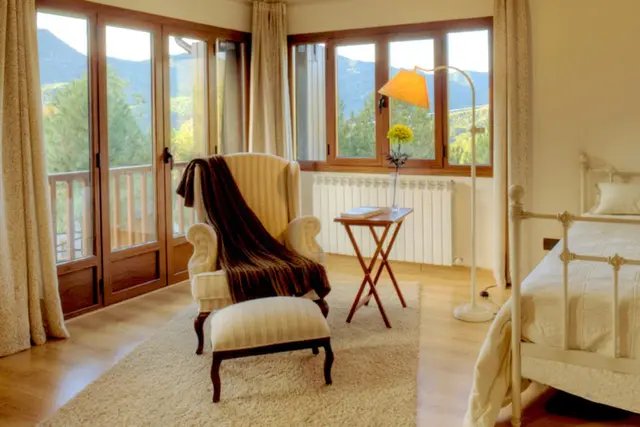 Interior de una habitación con una butaca y otomana en primer plano, una cama a la derecha, una mesa auxiliar