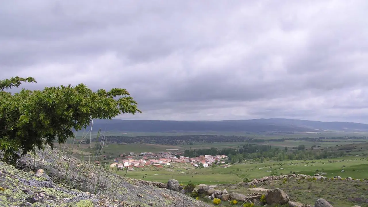 Vista panorámica de un pueblo en un valle rodeado de campos verdes y montañas lejanas, tomada desde un terreno rocoso
