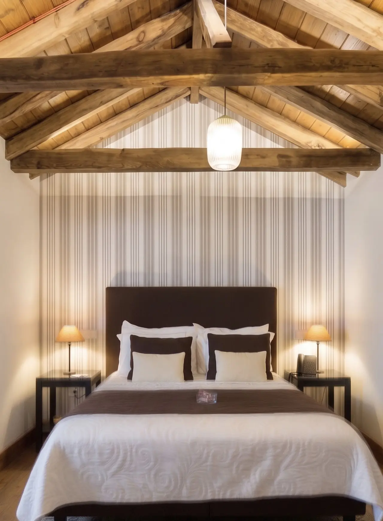Habitación con cama doble grande vestida en blanco y marrón, cabecero oscuro, dos mesitas de noche con lámparas