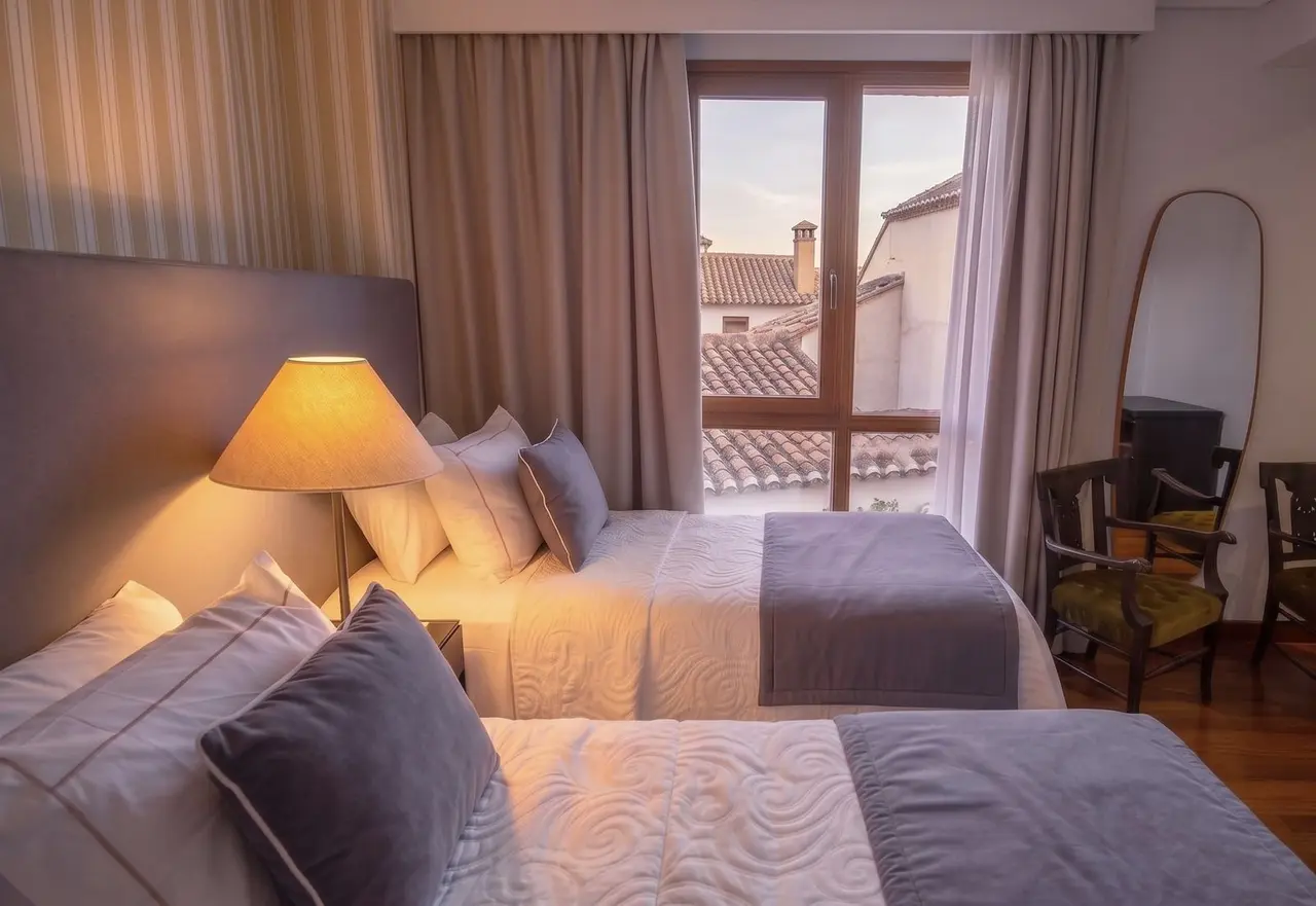 Habitación de hotel con dos camas grandes vestidas en tonos grises y blancos, cabecero oscuro, lámpara de mesita