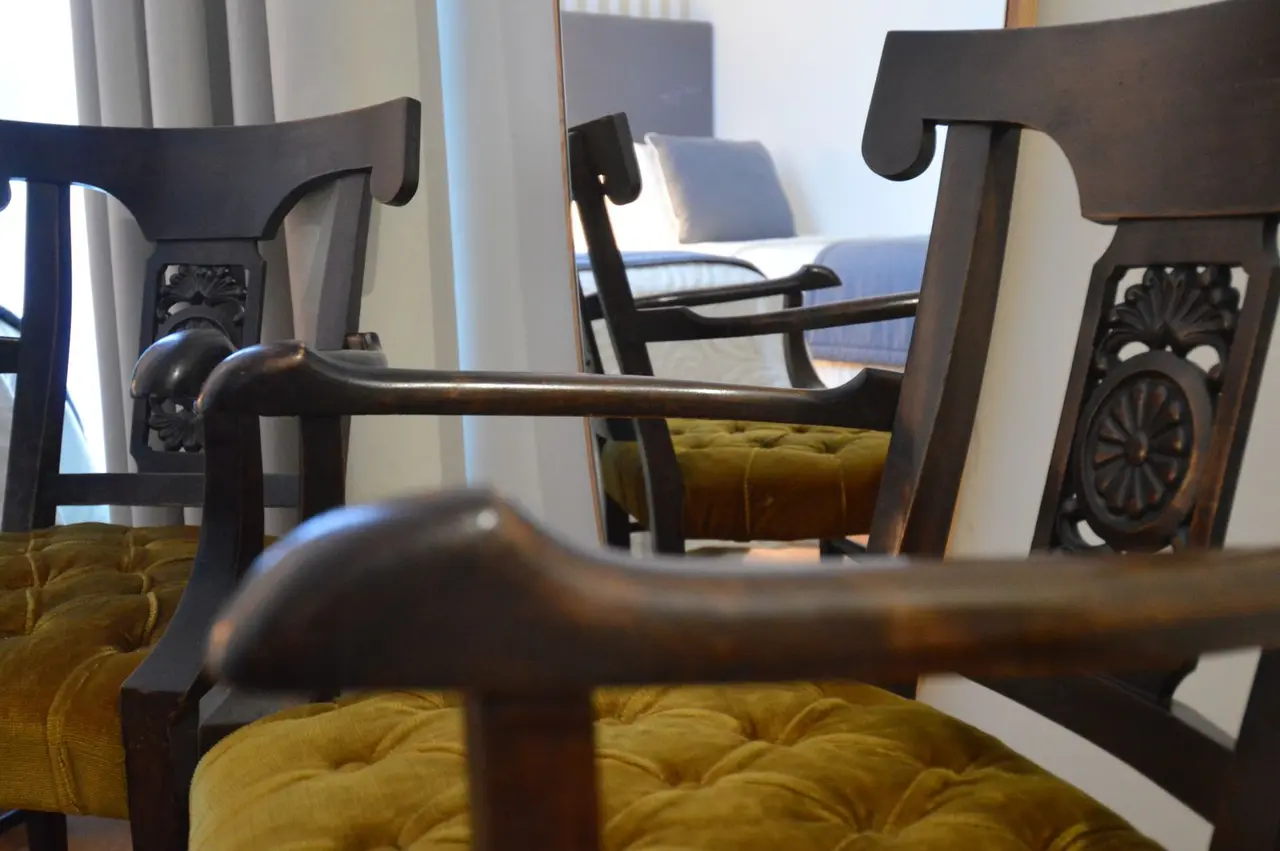 Sillas de madera oscura con tapizado amarillo mostaza en primer plano, reflejando en un espejo una cama con almohadas
