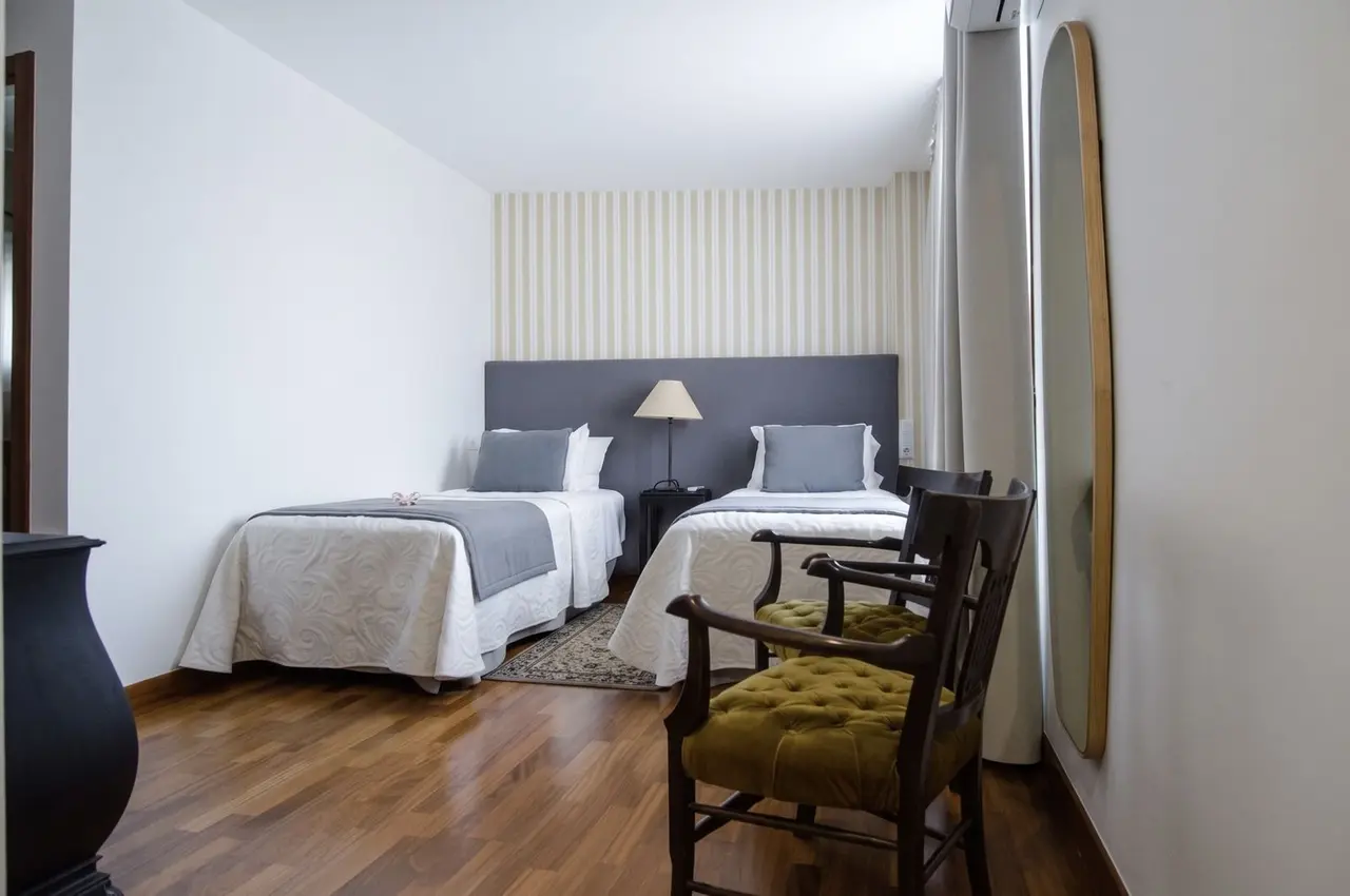 Habitación con dos camas individuales, cabeceros grises, sábanas blancas y una silla de madera con cojín verde
