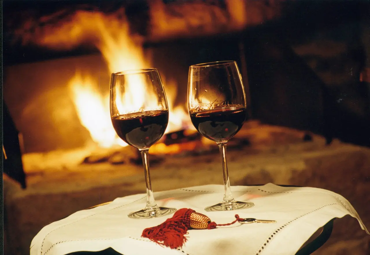 Dos copas de vino tinto sobre una mesa con mantel blanco frente a una chimenea encendida.