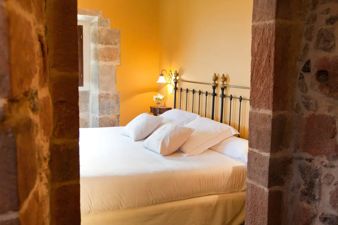 Vista de una habitación con cama doble de sábanas blancas y cabecero de forja, enmarcada por paredes de piedra antigua.
