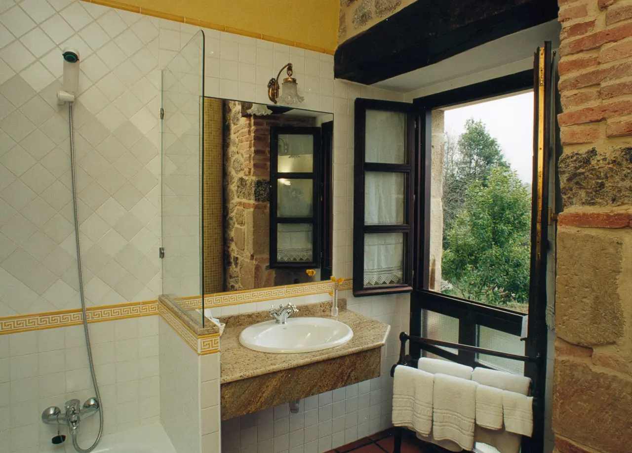 Baño con ducha, lavabo sobre encimera de piedra, espejo y ventana abierta con vistas a árboles y ladrillo visto.