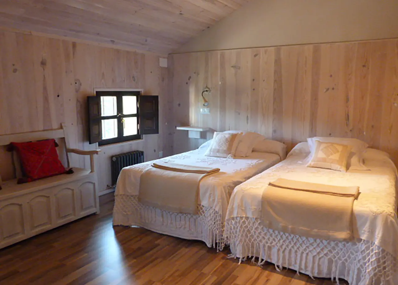 Habitación con dos camas individuales en un espacio con paredes y techo de madera clara, ventana con postigos