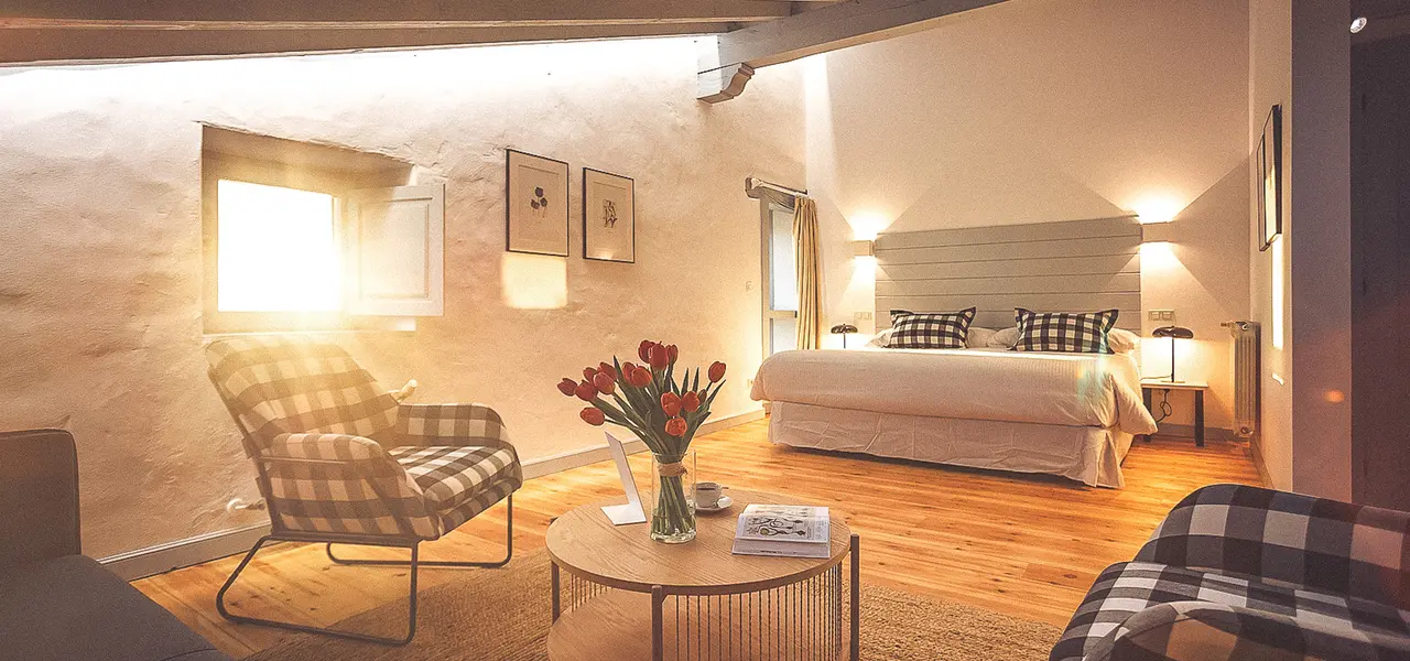 Habitación con paredes de piedra, suelo de madera, cama grande con cabecero blanco y dos sillones de cuadros