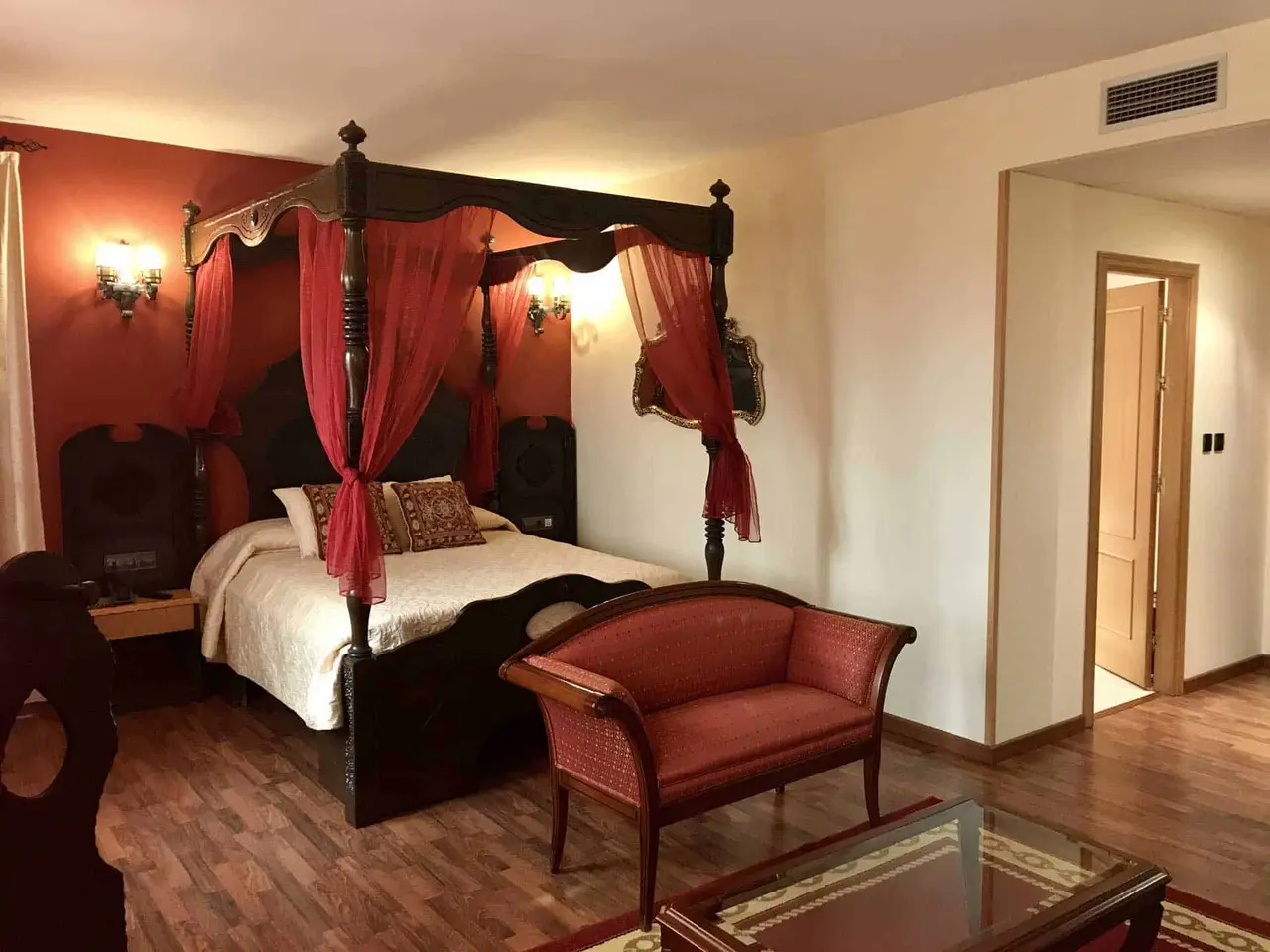 Vista de una suite de hotel con una gran cama con dosel de madera oscura y cortinas rojas, paredes en tonos rojos