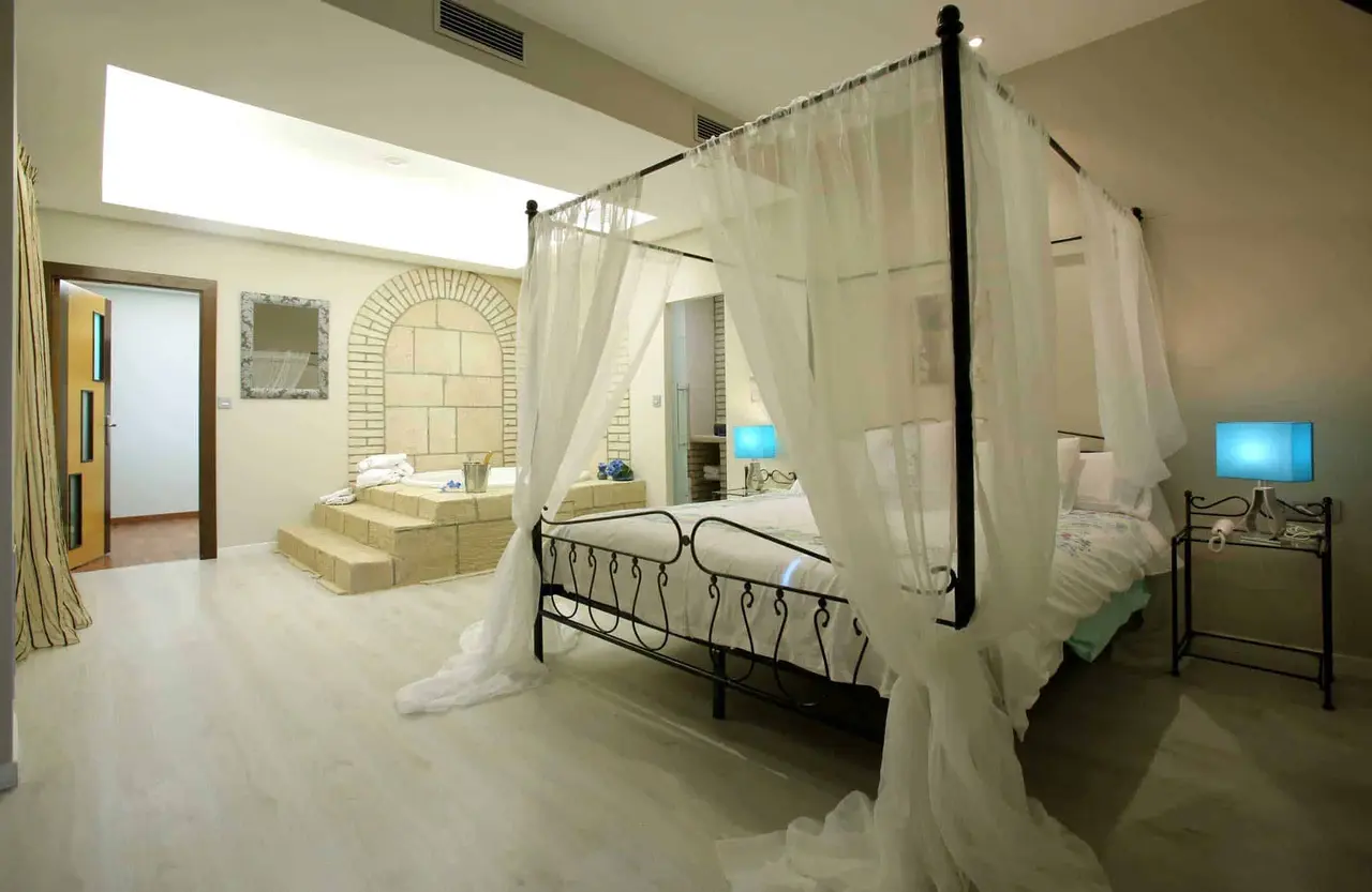 Vista de una suite con una gran cama con dosel de hierro forjado y cortinas blancas, suelo de baldosas claras