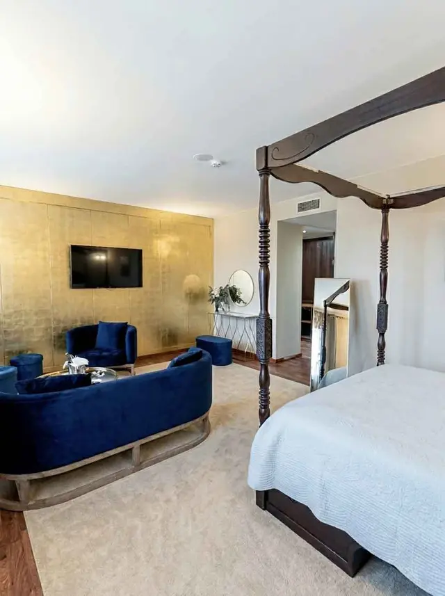 Vista de una suite de hotel con una gran cama de dosel de madera oscura, un sofá curvo azul en el centro