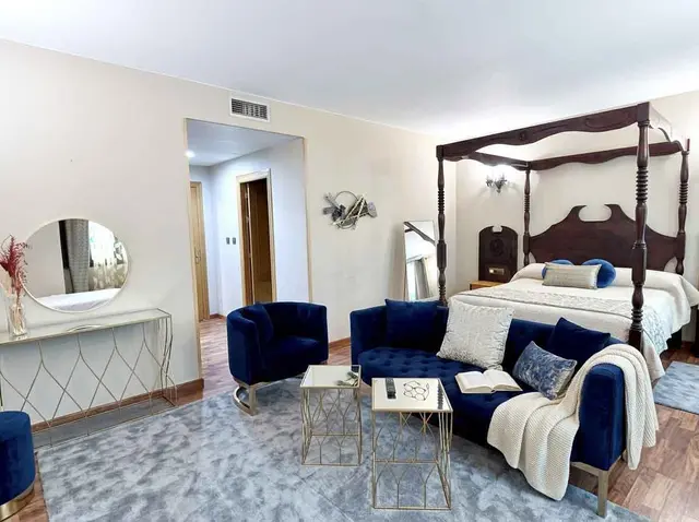 Vista de una suite de hotel con una gran cama con dosel de madera oscura, un sofá azul en forma de L, sillón azul