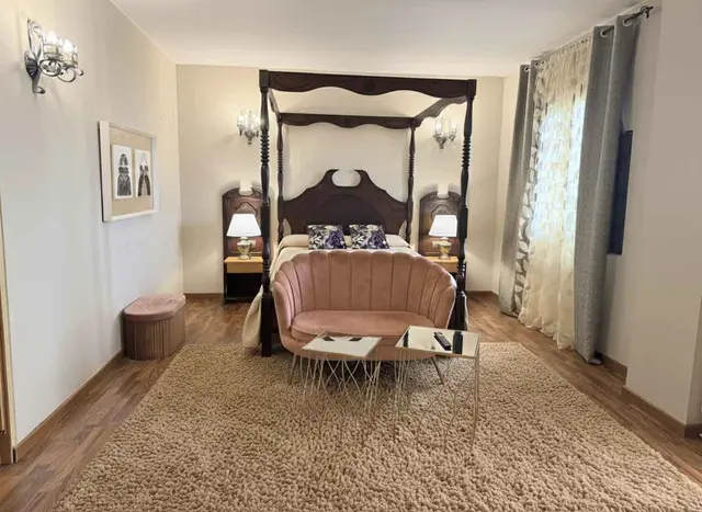 Vista de una suite de hotel con una gran cama de dosel de madera oscura, dos mesitas de noche con lámparas, un sofá