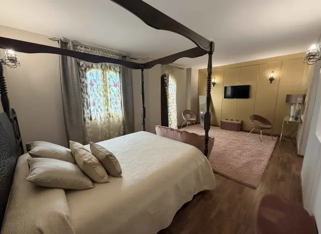 Vista de una suite de hotel con una gran cama de dosel con dosel de madera oscura y ropa de cama beige, alfombra rosa