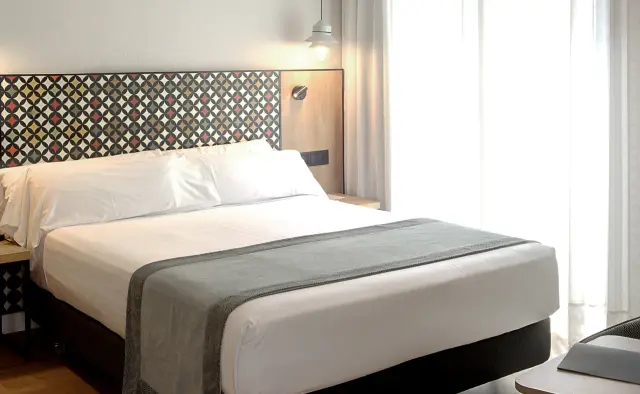 Habitación junior suite con cama king size vestida de blanco, cabecero decorado con patrón geométrico de mosaico
