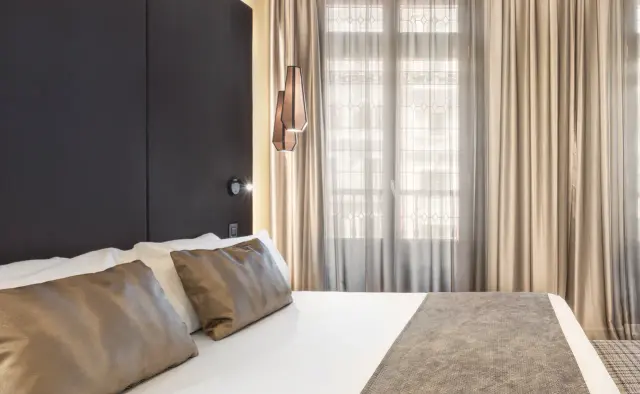 Interior de una habitación de hotel con cama grande vestida con sábanas blancas y cojines en tonos beige y marrones