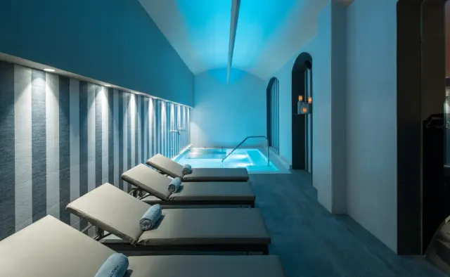 Interior de un spa con piscina iluminada en el fondo, varias tumbonas alineadas en primer plano sobre suelo de madera