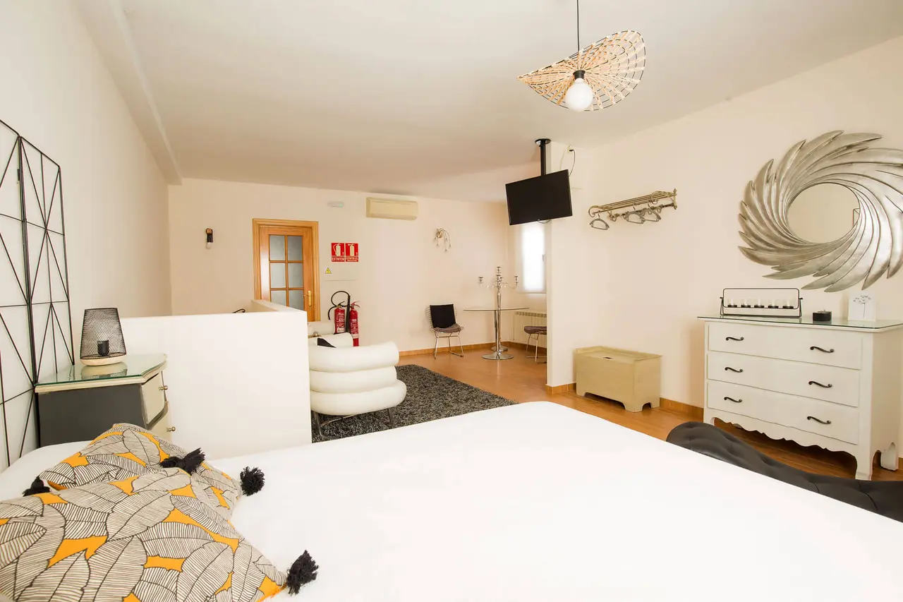 Habitación de alojamiento con cama grande en primer plano, sofá blanco, cómoda blanca, televisor montado en la pared