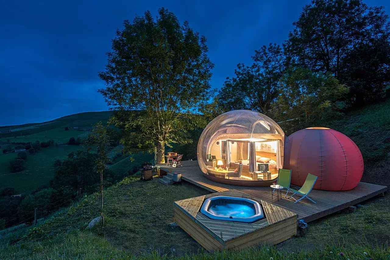 Jacuzzi privado exterior iluminado en una terraza de madera junto a una burbuja transparente en un paisaje rural