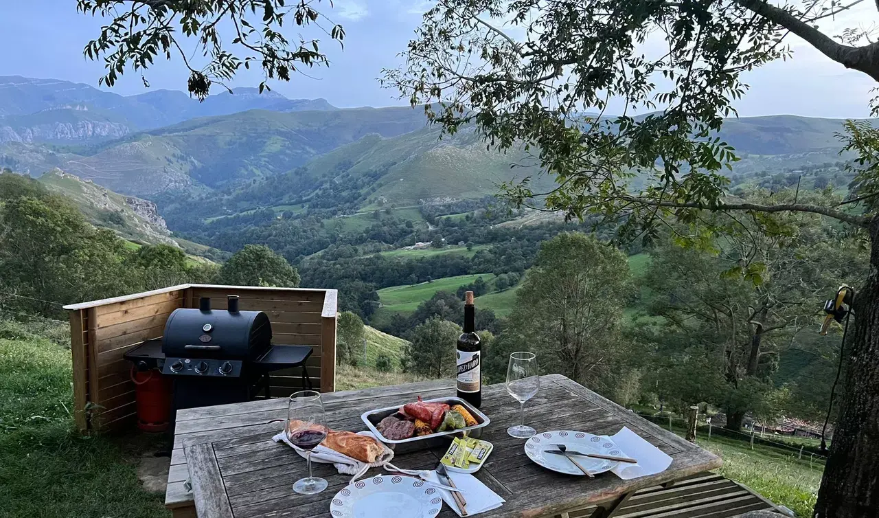 Mesa de madera con barbacoa, vino y comida preparada frente a un extenso paisaje de montañas y valles verdes.