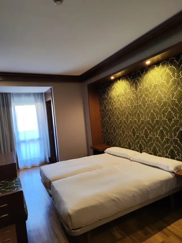 Habitación de hotel con dos camas individuales unidas, cabecero iluminado con papel pintado verde y blanco, mesitas de n
