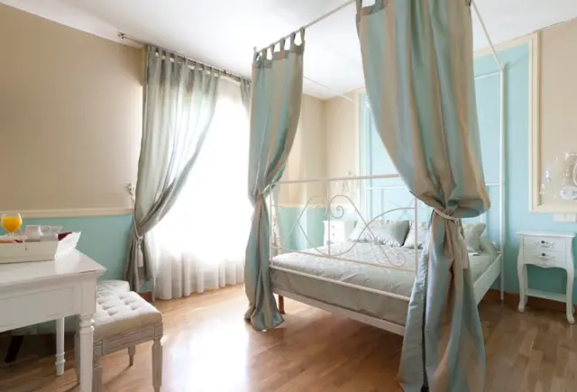 Habitación de hotel con cama de dosel con cortinas azules, escritorio blanco con banqueta, ventana con cortinas