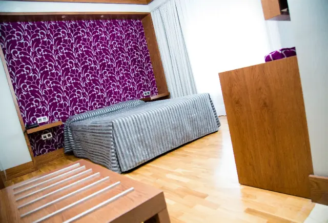Habitación de hotel con cama doble cubierta por una colcha a rayas grises y blancas, cabecero con papel pintado morado