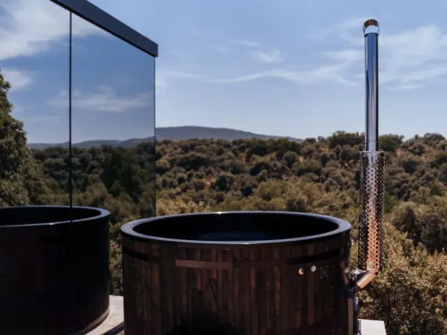 Jacuzzi nórdico de madera con chimenea en terraza frente a un bosque de montañas.