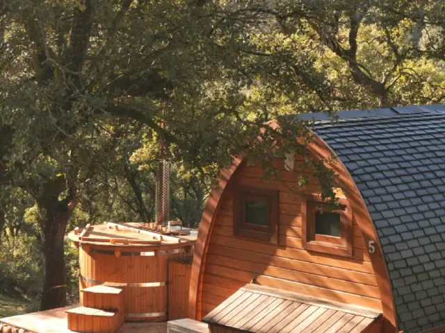 Jacuzzi nórdico de madera con chimenea en terraza junto a cabaña de madera rodeada de árboles.